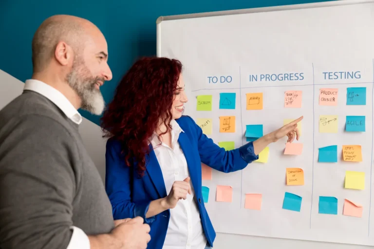Two colleagues discussing coloured sticky notes on a whiteboard task board