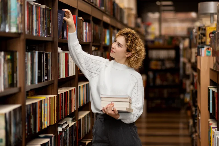 Librarian reaching for a book on a tall wooden shelf, holding a stack