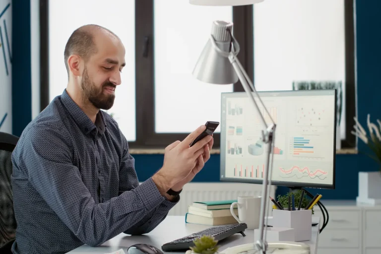 Man at an office desk checking his phone beside a monitor showing charts
