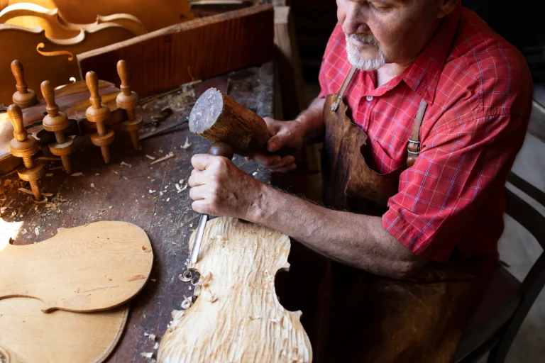 Older craftsman carving a violin scroll with chisel and mallet at a workbench