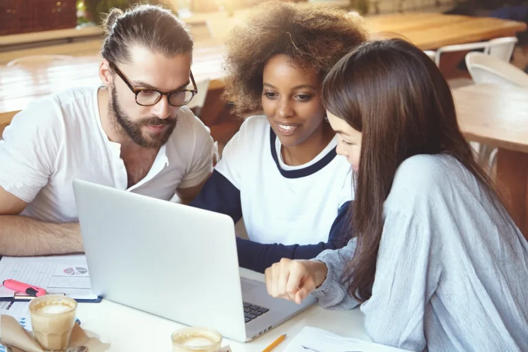 Three colleagues leaning in around a laptop at a cafe table